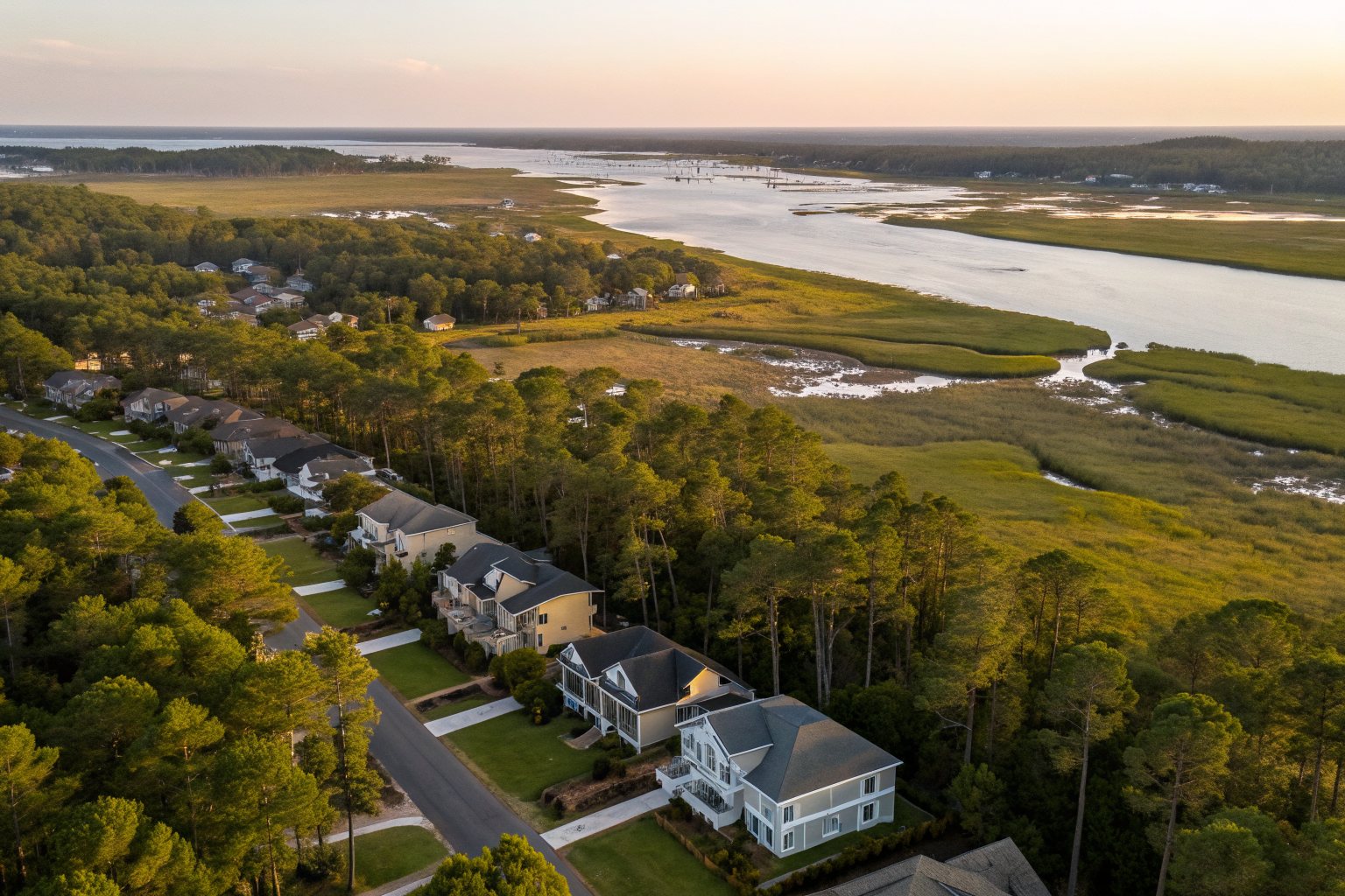 Aerial view of shingle-roofed homes along a pine-lined street beside a tidal salt marsh and a wide waterway