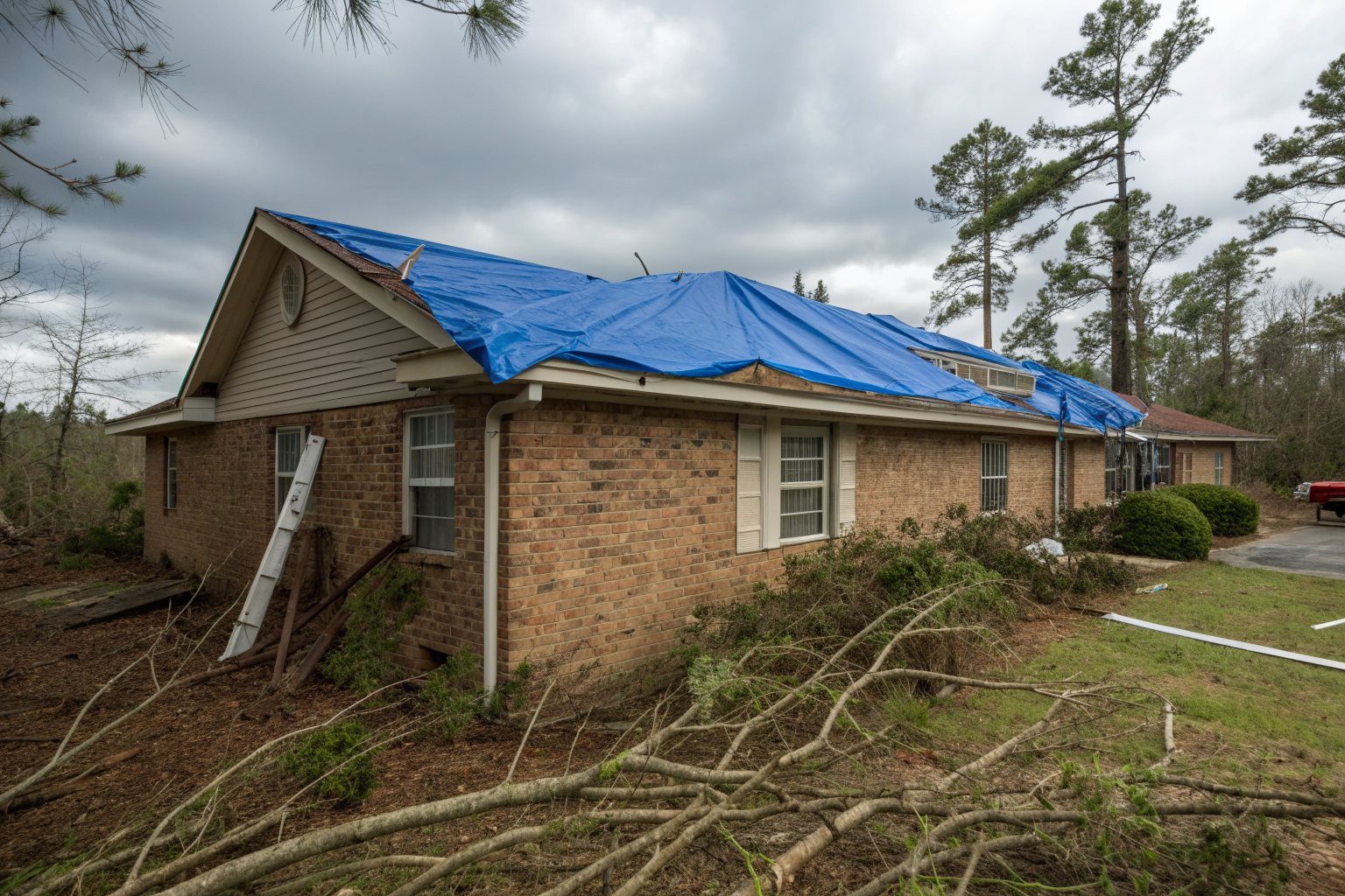 A blue tarp over the roof of a tan brick ranch house, with fallen pine branches in the yard